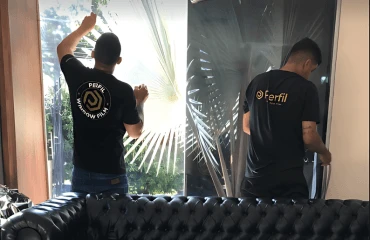 Dois instaladores da Perfil Window Film, de uniforme, aplicando película em um vidro de sala